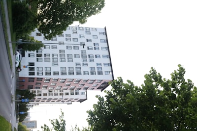 A multi-story residential or office building with a white and brick exterior, featuring numerous windows in a geometric pattern. The structure stands adjacent to a street where a parked vehicle and traffic signs are visible. Green leafy trees frame the scene, hinting at an urban environment with natural elements.