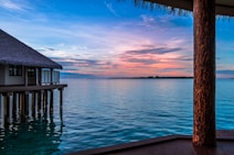 A serene view of a tropical ocean, featuring a thatched-roof bungalow on stilts over the water. The sky displays a beautiful gradient from blue to pink, reflecting gently on the still ocean surface. A wooden deck and column are visible in the foreground, adding a rustic touch to the tranquil setting.