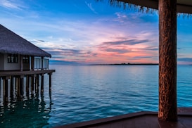 A serene view of a tropical ocean, featuring a thatched-roof bungalow on stilts over the water. The sky displays a beautiful gradient from blue to pink, reflecting gently on the still ocean surface. A wooden deck and column are visible in the foreground, adding a rustic touch to the tranquil setting.