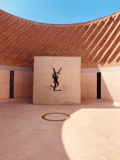 A modern architectural space with a circular courtyard and a central square wall featuring the letters 'YSL' mounted in contrast against a textured background. The surrounding high brick walls curve smoothly, creating an open airy space under a clear blue sky. Sunlight casts dynamic shadows on the ground.