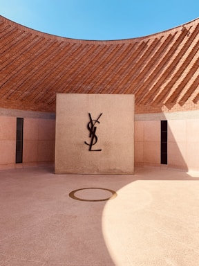 A modern architectural space with a circular courtyard and a central square wall featuring the letters 'YSL' mounted in contrast against a textured background. The surrounding high brick walls curve smoothly, creating an open airy space under a clear blue sky. Sunlight casts dynamic shadows on the ground.