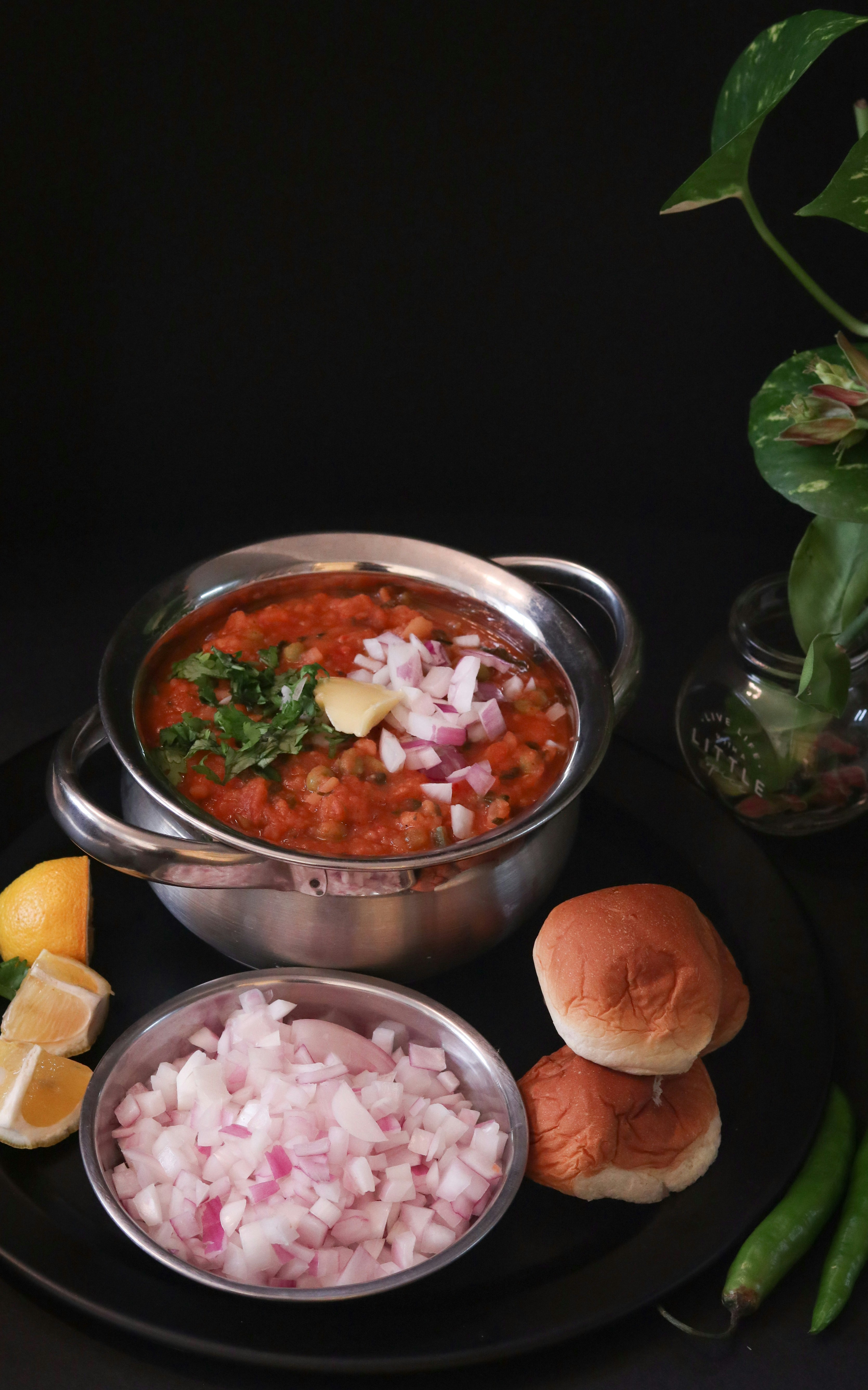 Pav bhaji topped with butter, onion and coriander in a steel pot, beside two pav buns, lemon wedges and a bowl of chopped onion