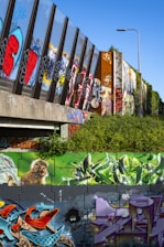 A vibrant display of street art and graffiti on a series of walls and vertical structures, showcasing various colorful designs, patterns, and text. A mural of a monkey is prominently featured alongside abstract shapes and vibrant lettering. The graffiti includes bold colors like red, blue, purple, and green on a background of sometimes grey and black walls. A street lamp and some greenery are visible, highlighting the urban environment.