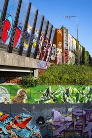 A vibrant display of street art and graffiti on a series of walls and vertical structures, showcasing various colorful designs, patterns, and text. A mural of a monkey is prominently featured alongside abstract shapes and vibrant lettering. The graffiti includes bold colors like red, blue, purple, and green on a background of sometimes grey and black walls. A street lamp and some greenery are visible, highlighting the urban environment.