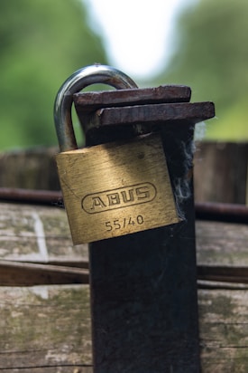 A close-up of a brass padlock with the brand name 'ABUS' and numbers '55/40' engraved on it. The lock is attached to an old, rusted metal latch on a wooden surface. The background is blurred, showing shades of green, suggesting a natural outdoor setting.