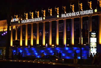 An illuminated building at night with tall columns and a row of statues holding bows on top. The facade is lit with warm yellow and blue lights, highlighting the signage reading 'PALACIO DE CONGRESOS'. A digital sign for the Hard Rock Cafe is visible, and several people and vehicles are seen in the foreground.