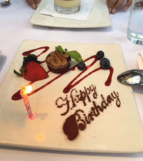 A square white plate features a birthday dessert with 'Happy Birthday' written in cocoa powder. There is a small chocolate tart topped with whipped cream and garnished with a strawberry, blueberries, and a mint leaf. Red syrup is artistically drizzled across the plate. A lit birthday candle is placed beside the dessert. Additionally, the background shows part of another plate and a person's hand.