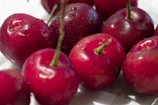 Several vibrant red cherries with green stems are closely arranged on a white surface. The cherries have a glossy sheen, suggesting they are fresh and recently washed. Some droplets of water are visible on their skins, enhancing their freshness and appeal.