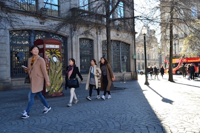 A group of four people is walking on a cobblestone street. They are dressed in winter clothing and appear to be sightseeing in an urban area. On the left side, there is a telephone booth decorated with colorful graffiti. In the background, a red open-top tour bus with passengers is parked. The scene is set in front of a historic building with large, ornate windows. The sunlight casts long shadows on the street, and there are a few trees with bare branches.