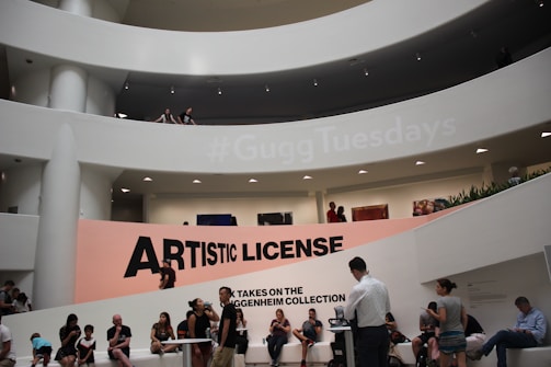 A modern architectural interior with a large, curved atrium and multiple people scattered throughout different levels. The prominent feature is a large sign reading 'Artistic License' positioned against a backdrop with a hashtag, '#GuggTuesdays', projected above. Several art pieces are visible on the walls, and people are both sitting and standing, engaged in various activities.