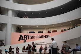 A modern architectural interior with a large, curved atrium and multiple people scattered throughout different levels. The prominent feature is a large sign reading 'Artistic License' positioned against a backdrop with a hashtag, '#GuggTuesdays', projected above. Several art pieces are visible on the walls, and people are both sitting and standing, engaged in various activities.