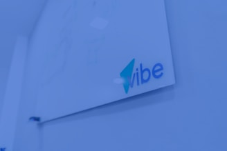 A whiteboard mounted on a wall with the word 'vibe' written in blue and teal colors. The surrounding environment is shaded in a blue tint, giving a modern and tech-focused appearance.