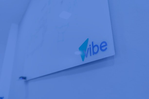 A whiteboard mounted on a wall with the word 'vibe' written in blue and teal colors. The surrounding environment is shaded in a blue tint, giving a modern and tech-focused appearance.