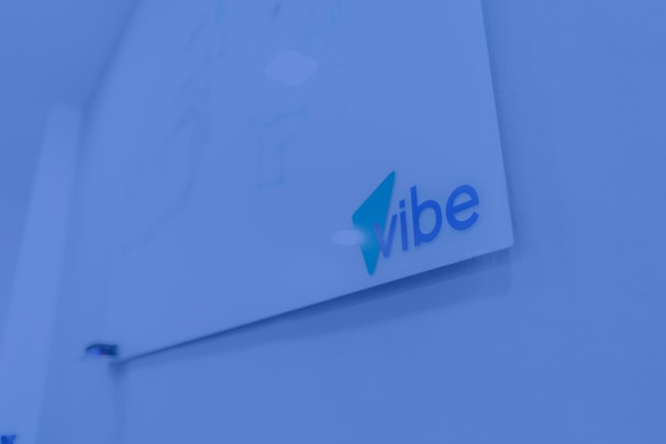 A whiteboard mounted on a wall with the word 'vibe' written in blue and teal colors. The surrounding environment is shaded in a blue tint, giving a modern and tech-focused appearance.