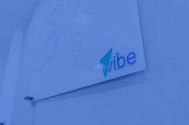 A whiteboard mounted on a wall with the word 'vibe' written in blue and teal colors. The surrounding environment is shaded in a blue tint, giving a modern and tech-focused appearance.