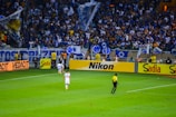 A crowded stadium with enthusiastic fans cheering, many of whom are wearing blue jerseys and holding flags. On the field, a referee in a yellow and black uniform is near the sideline, observing the match. Advertising boards for various brands like Nikon and Sadia line the edge of the field, while a player in white uniform walks nearby.