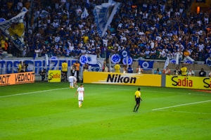A crowded stadium with enthusiastic fans cheering, many of whom are wearing blue jerseys and holding flags. On the field, a referee in a yellow and black uniform is near the sideline, observing the match. Advertising boards for various brands like Nikon and Sadia line the edge of the field, while a player in white uniform walks nearby.