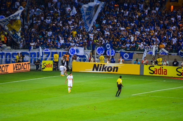 A crowded stadium with enthusiastic fans cheering, many of whom are wearing blue jerseys and holding flags. On the field, a referee in a yellow and black uniform is near the sideline, observing the match. Advertising boards for various brands like Nikon and Sadia line the edge of the field, while a player in white uniform walks nearby.
