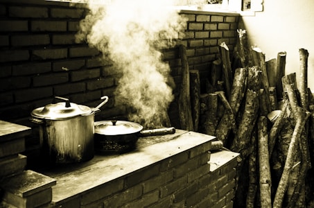 A rustic outdoor cooking area with a large metal pot and a pan sitting on a brick stove. Steam is rising from the pan, creating a hazy atmosphere. A stack of cut firewood is arranged neatly against the brick wall, suggesting a traditional cooking method.