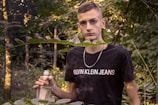 A young person is standing in a forested area, surrounded by green foliage. They are holding an empty glass bottle and wearing a black t-shirt with a visible brand logo. A silver chain necklace hangs around their neck, and the background is filled with trees and leaves.