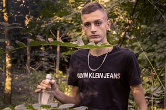 A young person is standing in a forested area, surrounded by green foliage. They are holding an empty glass bottle and wearing a black t-shirt with a visible brand logo. A silver chain necklace hangs around their neck, and the background is filled with trees and leaves.