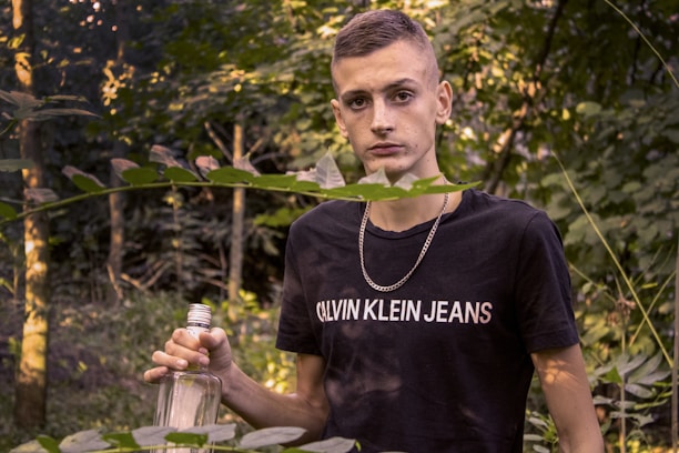 A young person is standing in a forested area, surrounded by green foliage. They are holding an empty glass bottle and wearing a black t-shirt with a visible brand logo. A silver chain necklace hangs around their neck, and the background is filled with trees and leaves.