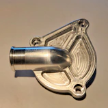 A close-up view of a shiny, metallic machined object with a cylindrical protrusion and intricate patterns. The object has a reflective surface, and the background appears to be a plain, light-colored surface.