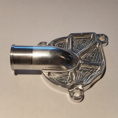 A metallic, intricately machined component with a tubular protrusion and detailed geometric patterns on its surface, likely a part for mechanical or industrial use.