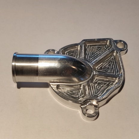A metallic, intricately machined component with a tubular protrusion and detailed geometric patterns on its surface, likely a part for mechanical or industrial use.