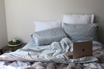 A cozy bed with rumpled gray bedding and white pillows hosts an open laptop. A notebook lies beside it, suggesting a relaxing or work-from-home setup. Next to the bed, a small side table holds a green plant and a candle.