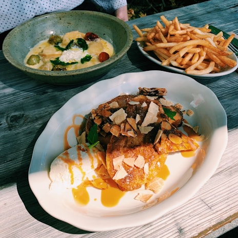 A rustic wooden table is set with three dishes: a round, grey bowl filled with a creamy pasta dish topped with cherry tomatoes, spinach, and cheese, a plate of golden crispy French fries, and a main dish of what appears to be French toast garnished with almond slices, caramel sauce, and a scoop of vanilla ice cream.