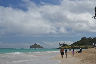 A beach scene with a family walking along the shoreline. The sky is partly cloudy with a picturesque island in the background. The ocean water is a clear turquoise, and the beach sand is light-colored. People are scattered across the beach, some walking and others sitting, enjoying the seaside view. There are a few houses and palm trees visible on a hill further away.