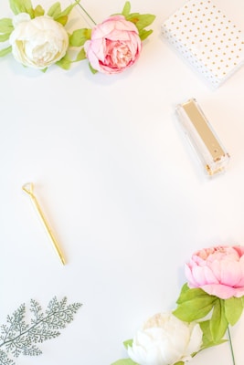 A minimalistic and aesthetically pleasing flatlay featuring a few peony flowers with white and pink petals, green leaves, a golden pen, a decorative fern-like leaf, a white box with golden dots, and a clear stapler with a golden top. The items are arranged against a white background, creating an elegant and clean composition.