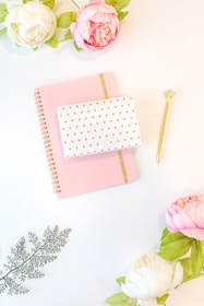 A flat lay featuring a pink spiral notebook with a gold elastic band, a white gift box with small gold hearts, and a gold pen with a crystal top. Surrounding these items are artificial flowers, including pink and white peonies, and a sage green fern leaf.