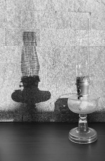 A vintage glass oil lamp stands on a dark table, casting a prominent shadow on a textured stone wall in the background. The intricate detailing on the lamp’s metal and glass surface is highlighted by the light.