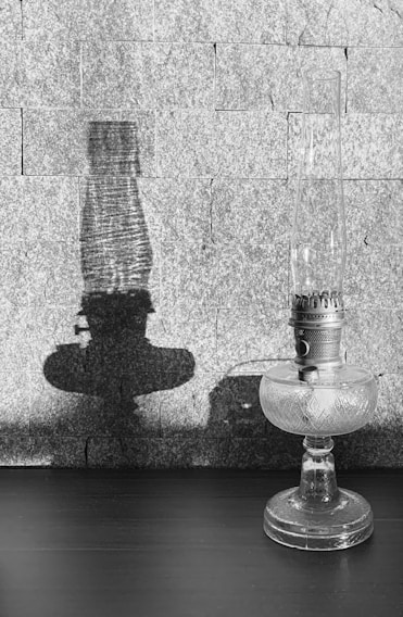 A vintage glass oil lamp stands on a dark table, casting a prominent shadow on a textured stone wall in the background. The intricate detailing on the lamp’s metal and glass surface is highlighted by the light.