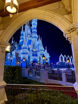 A fairy-tale castle is beautifully illuminated with sparkling blue and white lights against a dark night sky. The foreground features an arched stone entrance with vintage-style lanterns, and the castle is surrounded by ornate railings and lush greenery.