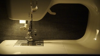 A close-up view of a sewing machine with its needle and presser foot visible. The machine is illuminated, casting light on its white surface, while the background remains dark.
