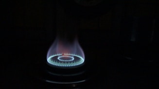 A gas stove burner is lit, emitting a bright blue flame arranged in a circular pattern. The flame subtly transitions from blue at the base to a hint of red at the tip, casting a soft glow in the surrounding darkness.