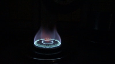 A gas stove burner is lit, emitting a bright blue flame arranged in a circular pattern. The flame subtly transitions from blue at the base to a hint of red at the tip, casting a soft glow in the surrounding darkness.