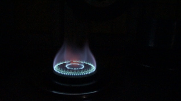 A gas stove burner is lit, emitting a bright blue flame arranged in a circular pattern. The flame subtly transitions from blue at the base to a hint of red at the tip, casting a soft glow in the surrounding darkness.