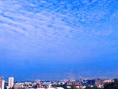 A cityscape with several buildings of varying heights, including a tall structure with the word 'METRO' prominently displayed. The sky is filled with scattered, fluffy clouds and has a gradient of blue hues.
