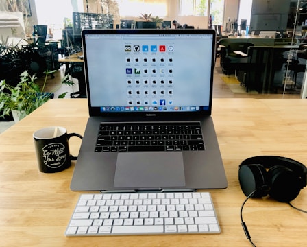 A wooden desk is equipped with a laptop displaying an array of application icons on the screen. To the left of the laptop is a black mug, and to the right is a set of black headphones. In front of the laptop, there is a wireless keyboard. The background reveals an office setting with plants and several workstations.