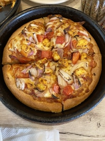 A pan pizza with a thick crust topped with chunks of paneer, tomato pieces, sliced onions, and a generous amount of melted cheese is placed in a black baking pan. The pizza appears to be freshly baked and is set on a wooden table with some spices visible in the background.