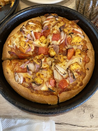 A pan pizza with a thick crust topped with chunks of paneer, tomato pieces, sliced onions, and a generous amount of melted cheese is placed in a black baking pan. The pizza appears to be freshly baked and is set on a wooden table with some spices visible in the background.