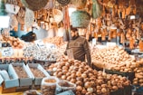 A bustling market stall filled with a variety of produce, including piles of onions, potatoes, and other vegetables. Numerous bottles and jars are neatly arranged in the foreground. The market is adorned with hanging baskets and cloves of garlic. A person wearing a sweater is examining the produce amidst the warm ambient light.