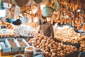 A bustling market stall filled with a variety of produce, including piles of onions, potatoes, and other vegetables. Numerous bottles and jars are neatly arranged in the foreground. The market is adorned with hanging baskets and cloves of garlic. A person wearing a sweater is examining the produce amidst the warm ambient light.