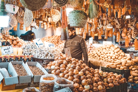 A bustling market stall filled with a variety of produce, including piles of onions, potatoes, and other vegetables. Numerous bottles and jars are neatly arranged in the foreground. The market is adorned with hanging baskets and cloves of garlic. A person wearing a sweater is examining the produce amidst the warm ambient light.