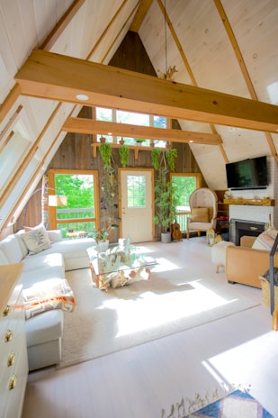 A cozy A-frame living room with high ceilings and wooden beams. Large windows allow natural light to fill the space, highlighting a mix of modern and rustic decor. There's a white sectional sofa, a wooden coffee table with artistic driftwood, and plants on the windowsill. A guitar rests against the wall, and a television is mounted above a white brick fireplace. A woven chair and a few decorative items complete the inviting atmosphere.