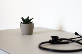 A small, dark green succulent plant in a beige pot is placed on a light-colored table. Nearby, a black stethoscope lies coiled, with part of it slightly out of focus. The background is plain and white, adding to the minimalist aesthetic.